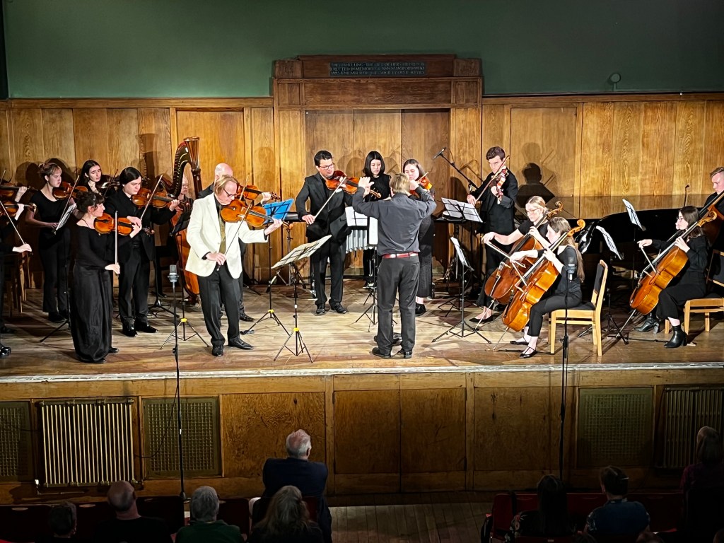 Viola player Richard Crabtree in white jacket, with a conductor and string players, harpist and vibraphone player