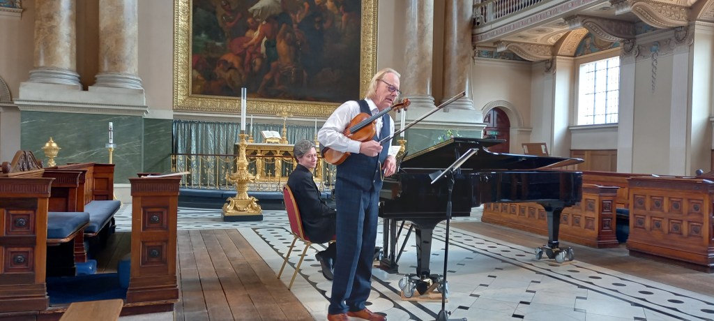 Male violist holding instrument and looking at music. Female pianist seated behind at grand piano. In an ornate chapel