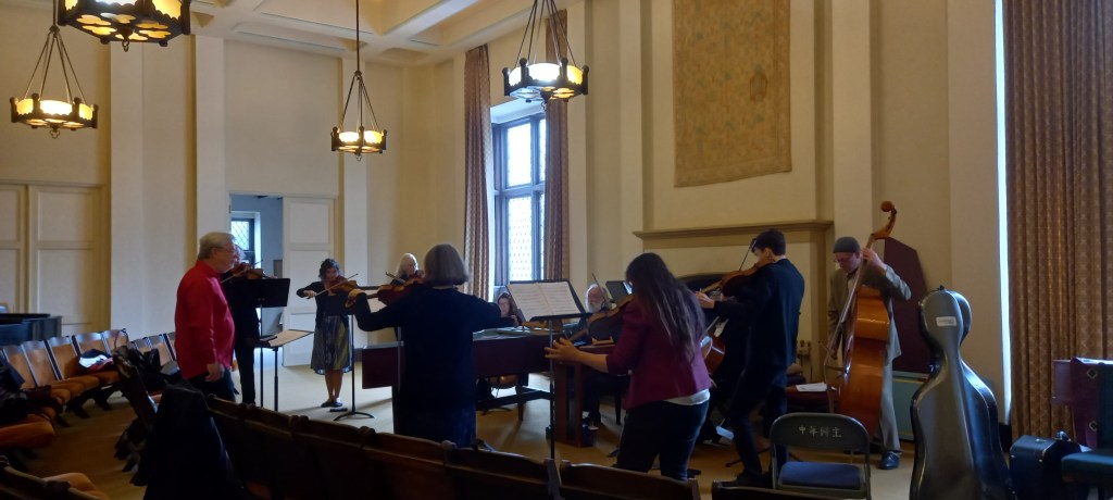 Group of musicians plus conductor rehearsing a piece in a room with windows and pictures