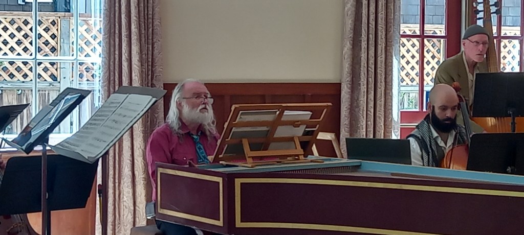 Harpsichordist, cellist and double bass player playing together in a room with windows behind
