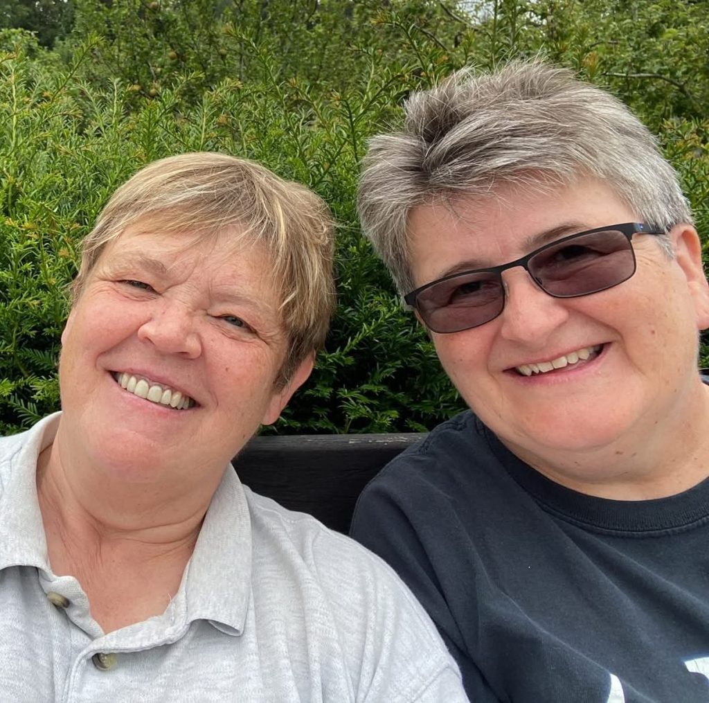 Selfie of two women seated on a bench, smiling at the camera, summer trees and bushed behind them, head shots.