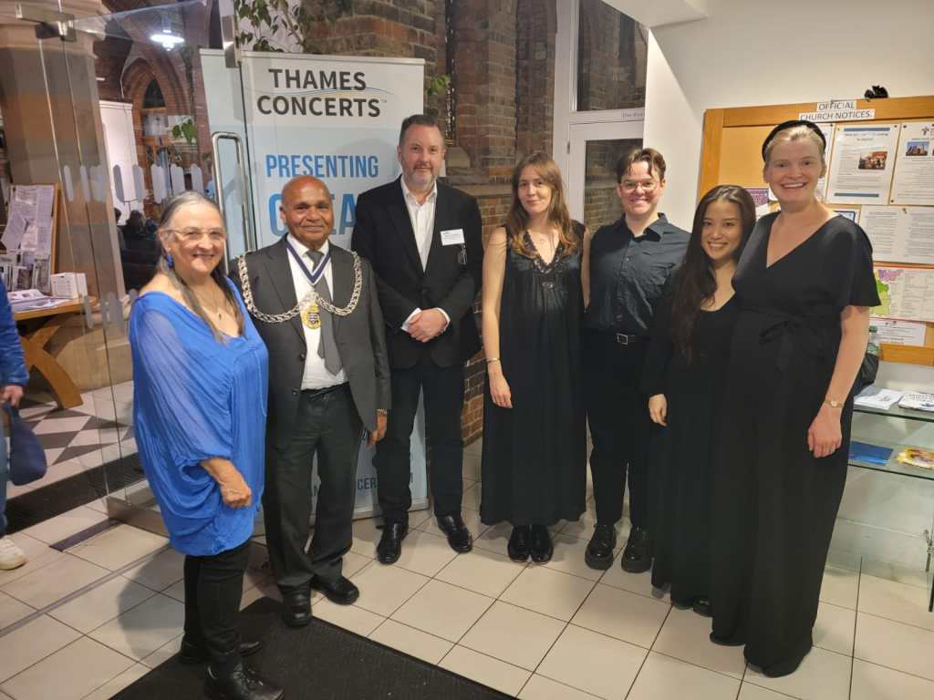 Seven people standing smiling at the camera, with Thames Concert banner behind them